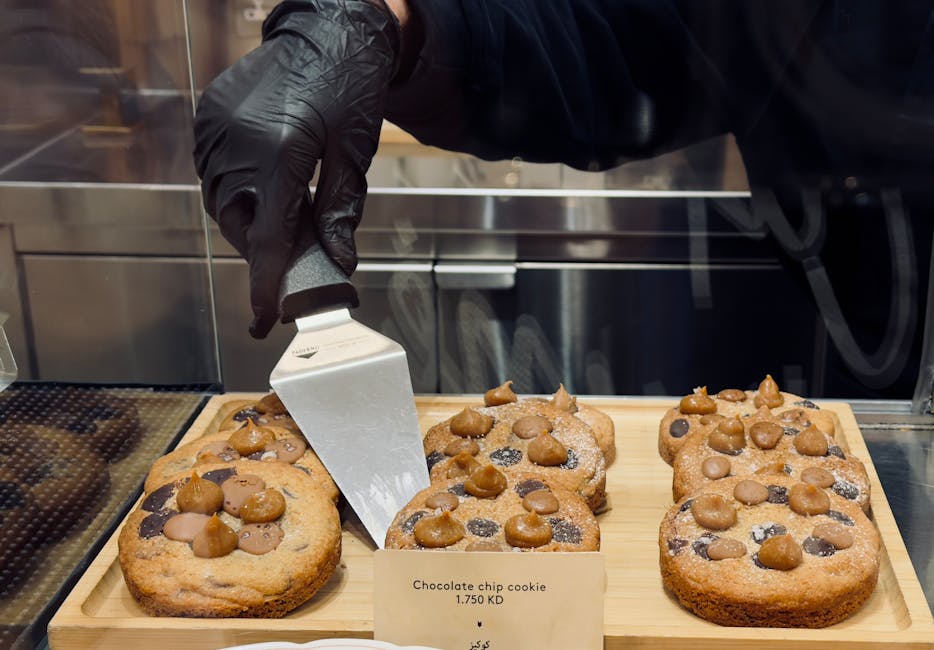A gloved hand serves chocolate chip cookies at a bakery counter displaying delicious baked goods.